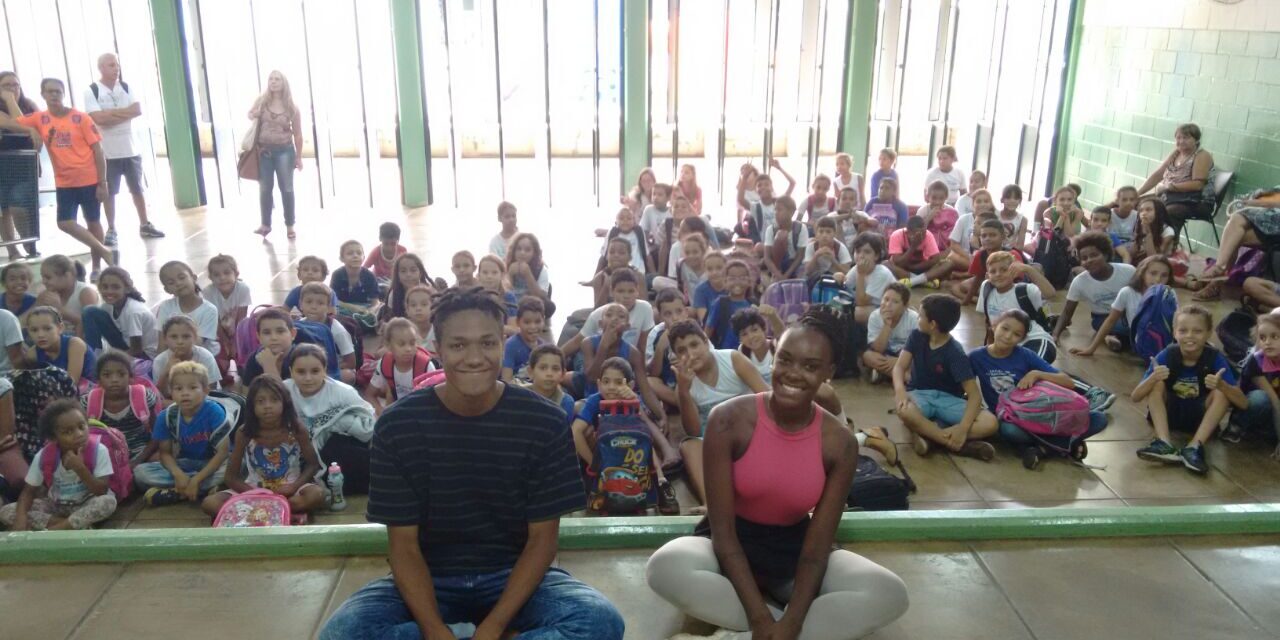 Dois instrutores posam com grande grupo de alunos sentados em sala de aula. Dois jovens sentados no chão, sorrindo, com um grande grupo de crianças sentadas atrás deles em uma sala iluminada.