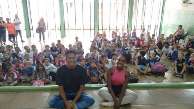 Dois instrutores posam com grande grupo de alunos sentados em sala de aula. Dois jovens sentados no chão, sorrindo, com um grande grupo de crianças sentadas atrás deles em uma sala iluminada.