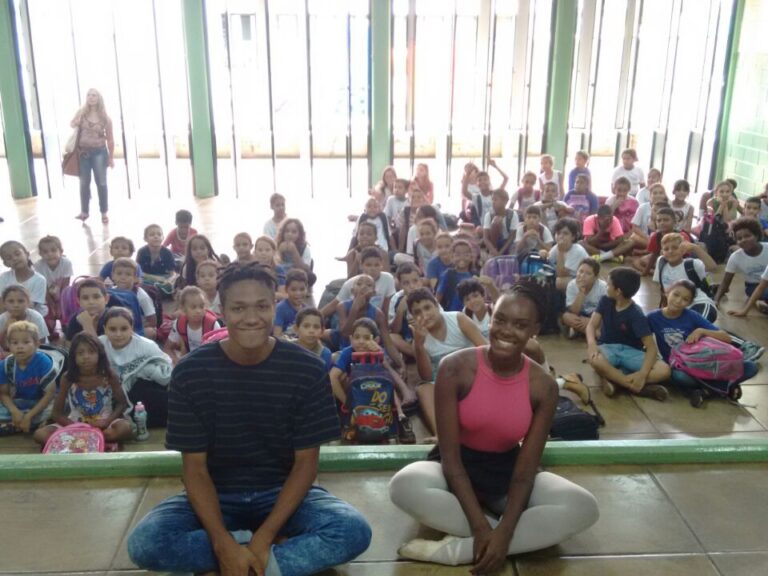 Dois instrutores posam com grande grupo de alunos sentados em sala de aula. Dois jovens sentados no chão, sorrindo, com um grande grupo de crianças sentadas atrás deles em uma sala iluminada.