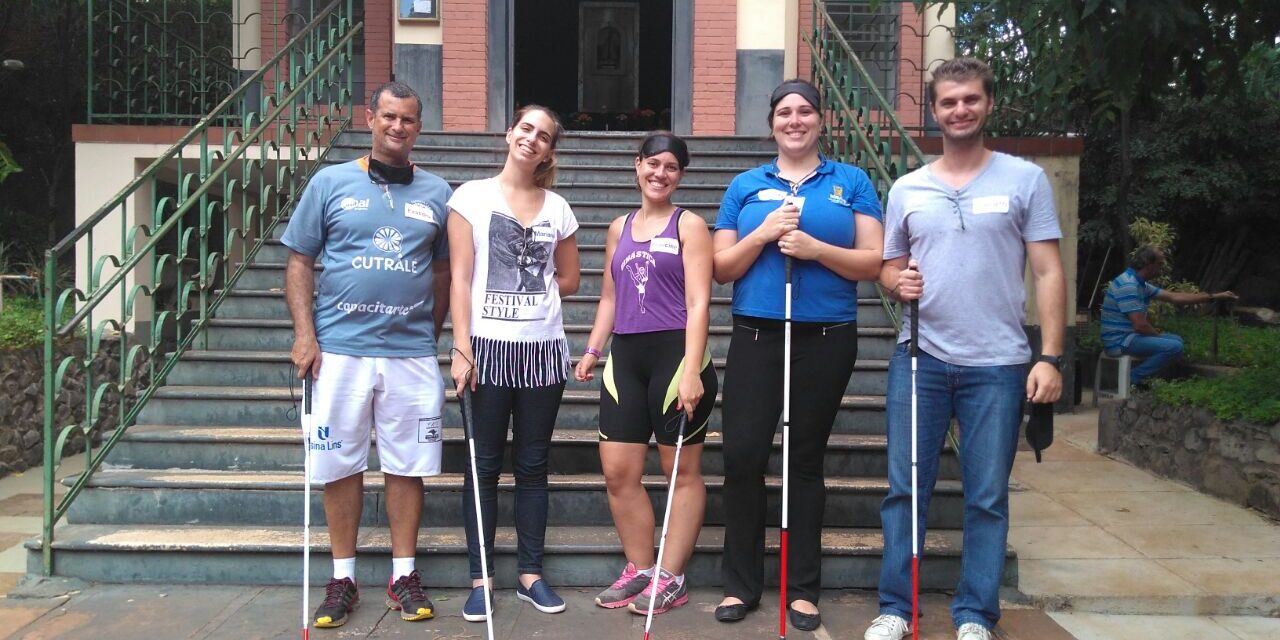 Grupo de apoio e inclusão posa com bengalas na escadaria. Cinco adultos, segurando bengalas brancas, posam na frente de uma escadaria de prédio com jardim ao lado.