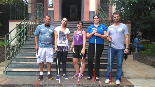 Grupo de apoio e inclusão posa com bengalas na escadaria. Cinco adultos, segurando bengalas brancas, posam na frente de uma escadaria de prédio com jardim ao lado.