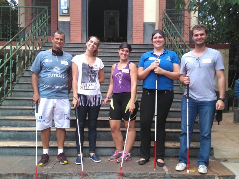 Grupo de apoio e inclusão posa com bengalas na escadaria. Cinco adultos, segurando bengalas brancas, posam na frente de uma escadaria de prédio com jardim ao lado.