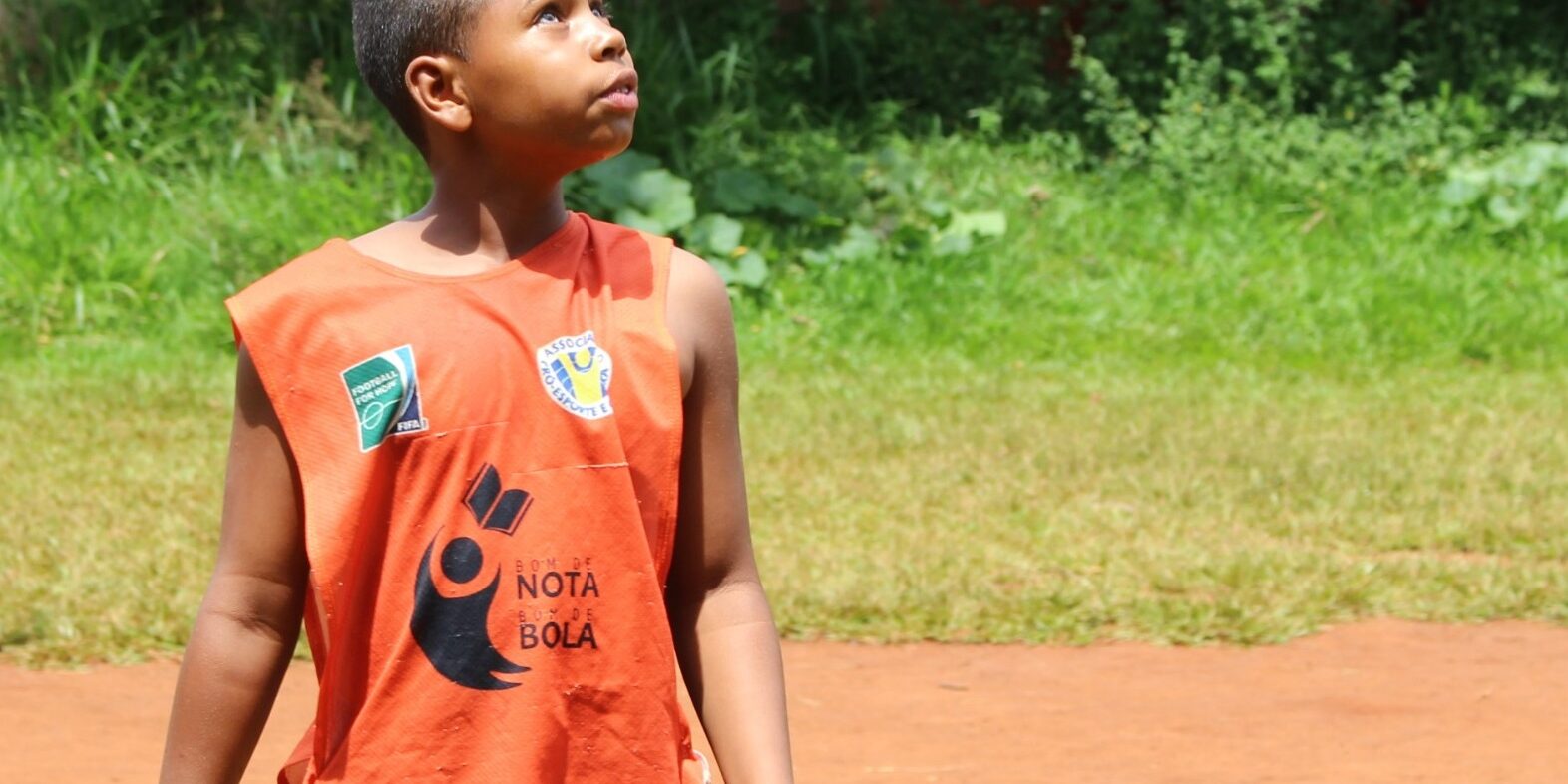 Aluno em quadra de terra olha para o céu. Menino de camiseta regata laranja "Nota 10 na Bola" em uma quadra de terra, olhando para cima.