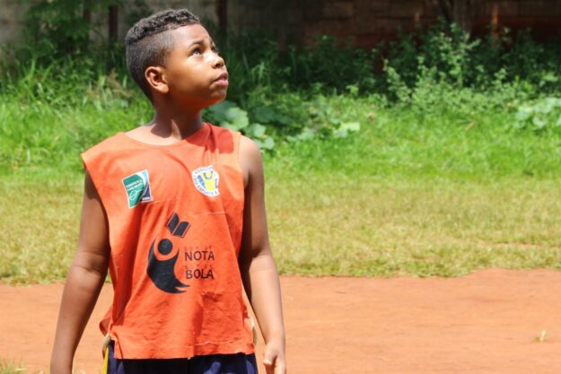 Aluno em quadra de terra olha para o céu. Menino de camiseta regata laranja "Nota 10 na Bola" em uma quadra de terra, olhando para cima.