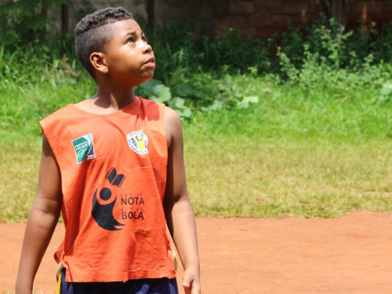 Aluno em quadra de terra olha para o céu. Menino de camiseta regata laranja "Nota 10 na Bola" em uma quadra de terra, olhando para cima.