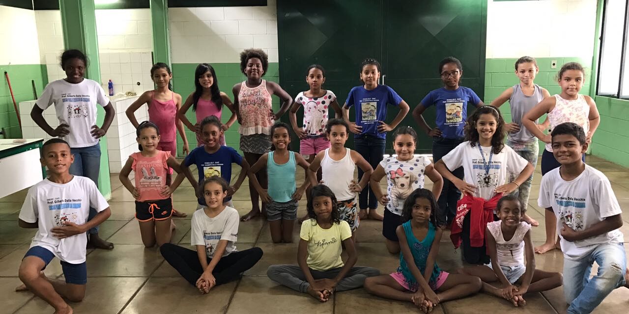Retrato de grupo de dança no local de ensaio. Grupo de crianças em trajes casuais posa em uma sala, algumas sentadas no chão e outras em pé atrás.