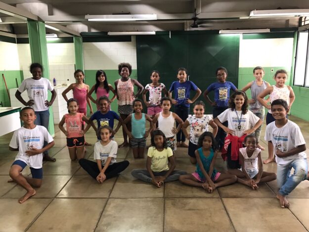 Retrato de grupo de dança no local de ensaio. Grupo de crianças em trajes casuais posa em uma sala, algumas sentadas no chão e outras em pé atrás.