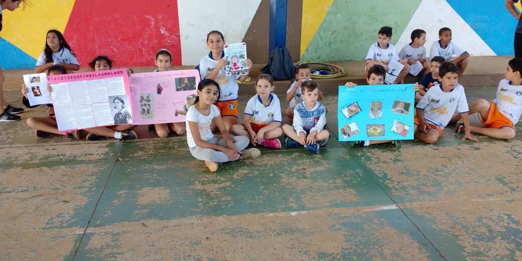 Estudantes exibem cartazes sentados em frente a mural colorido. Grupo de crianças sentadas no chão segurando cartazes e um livro, em frente a uma parede colorida.