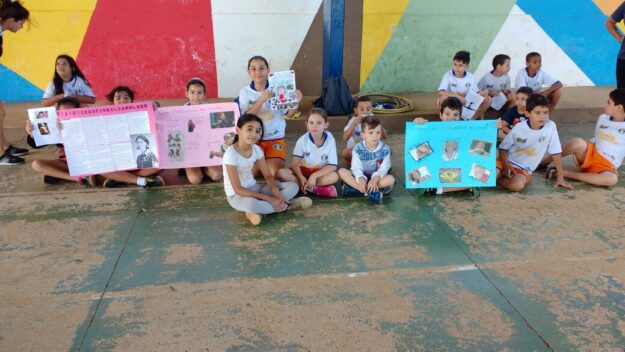 Estudantes exibem cartazes sentados em frente a mural colorido. Grupo de crianças sentadas no chão segurando cartazes e um livro, em frente a uma parede colorida.