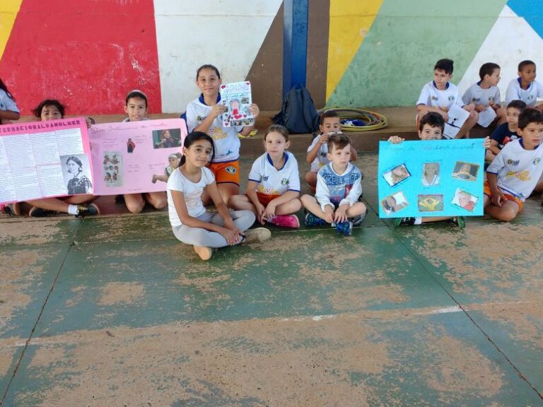 Estudantes exibem cartazes sentados em frente a mural colorido. Grupo de crianças sentadas no chão segurando cartazes e um livro, em frente a uma parede colorida.