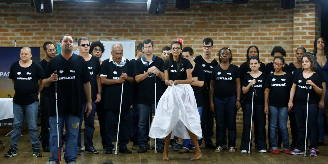 Apresentação artística de grupo de deficientes visuais. Grupo de deficientes visuais com camisetas pretas e bengalas em palco, enquanto uma mulher no centro dança.