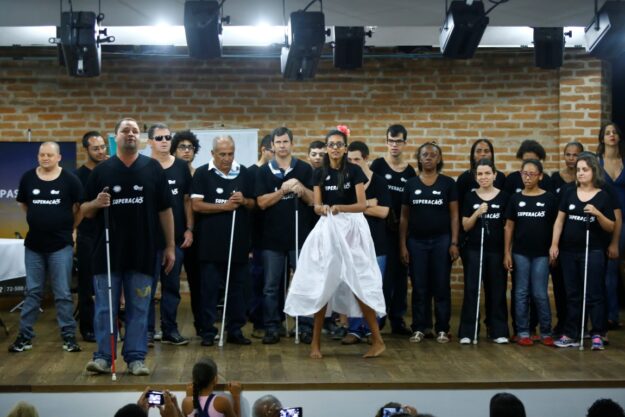 Apresentação artística de grupo de deficientes visuais. Grupo de deficientes visuais com camisetas pretas e bengalas em palco, enquanto uma mulher no centro dança.