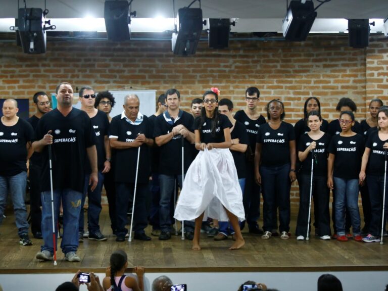 Apresentação artística de grupo de deficientes visuais. Grupo de deficientes visuais com camisetas pretas e bengalas em palco, enquanto uma mulher no centro dança.