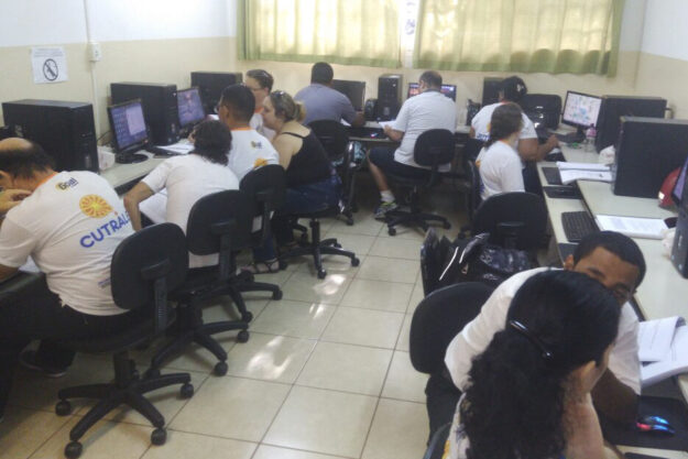 Aula de informática. Várias pessoas em uma sala de aula de informática, sentadas em cadeiras de rodinhas e olhando em computadores.