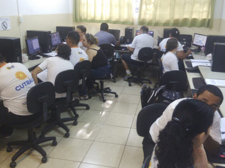 Aula de informática. Várias pessoas em uma sala de aula de informática, sentadas em cadeiras de rodinhas e olhando em computadores.
