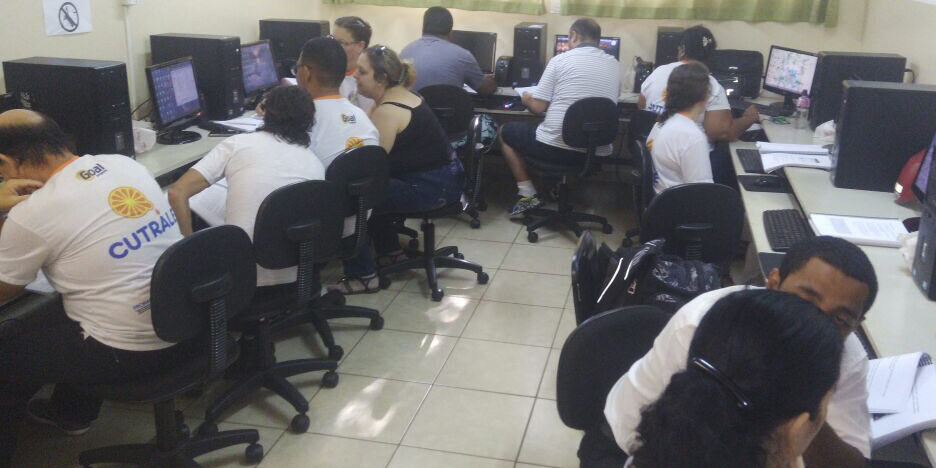 Aula de informática. Várias pessoas em uma sala de aula de informática, sentadas em cadeiras de rodinhas e olhando em computadores.