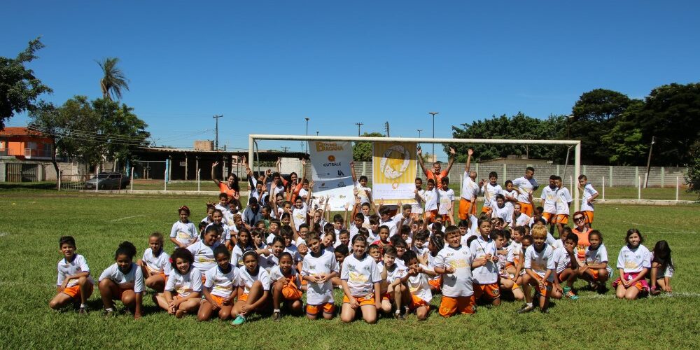 Foto de grupo de jovens alunos em campo de futebol sob céu azul. Grande grupo de crianças em uniformes brancos e laranjas posa em um campo de futebol, de joelhos e em pé, com um gol atrás.