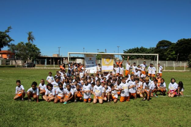 Foto de grupo de jovens alunos em campo de futebol sob céu azul. Grande grupo de crianças em uniformes brancos e laranjas posa em um campo de futebol, de joelhos e em pé, com um gol atrás.