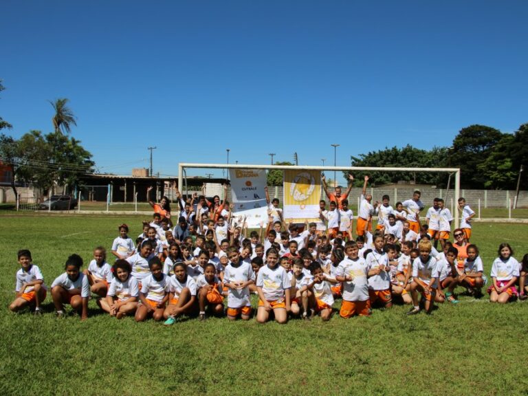 Foto de grupo de jovens alunos em campo de futebol sob céu azul. Grande grupo de crianças em uniformes brancos e laranjas posa em um campo de futebol, de joelhos e em pé, com um gol atrás.