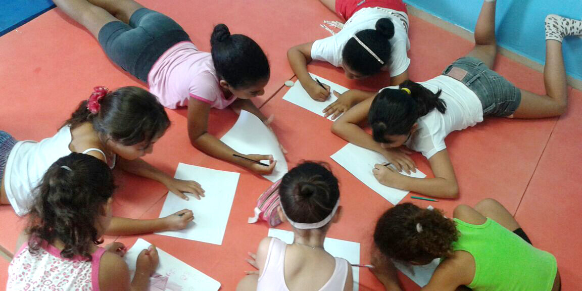 Aula de arte: Meninas desenham deitadas em grupo. Sete meninas, deitadas de bruços em um tapete vermelho, desenham em folhas de papel.