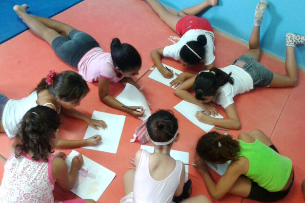 Sete meninas, deitadas de bruços em um tapete vermelho, desenham em folhas de papel.