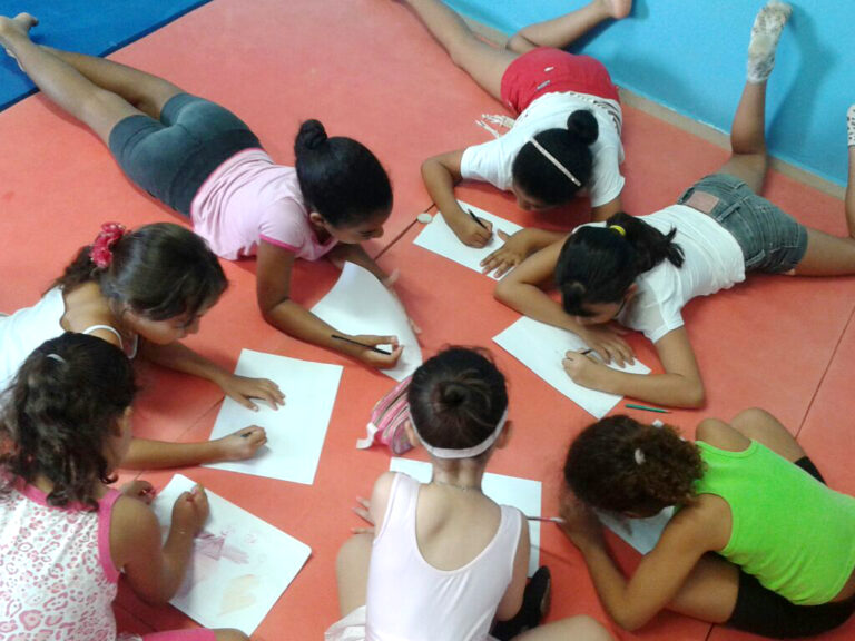 Aula de arte: Meninas desenham deitadas em grupo. Sete meninas, deitadas de bruços em um tapete vermelho, desenham em folhas de papel.