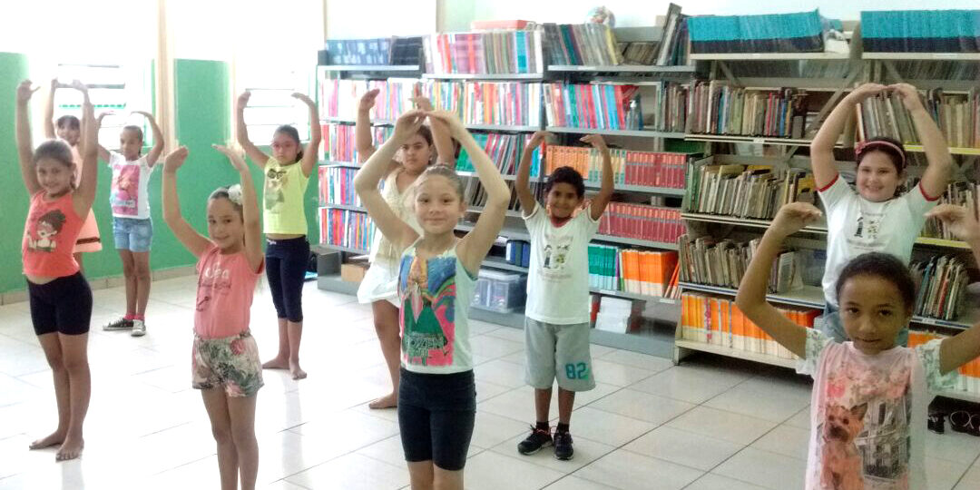 Crianças praticam movimentos em frente a estantes de livros. Grupo de crianças pratica movimentos de dança, com os braços erguidos em forma de círculo, em uma biblioteca com estantes cheias.