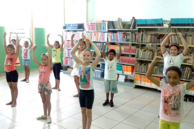 Crianças praticam movimentos em frente a estantes de livros. Grupo de crianças pratica movimentos de dança, com os braços erguidos em forma de círculo, em uma biblioteca com estantes cheias.