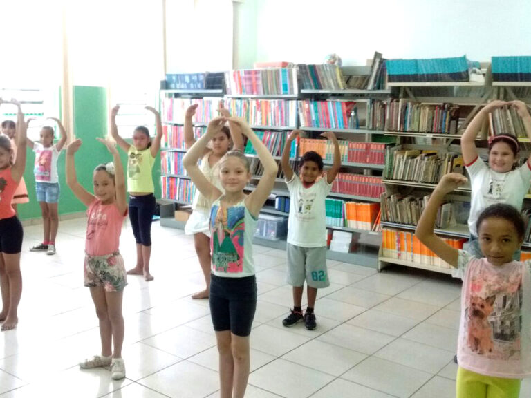 Crianças praticam movimentos em frente a estantes de livros. Grupo de crianças pratica movimentos de dança, com os braços erguidos em forma de círculo, em uma biblioteca com estantes cheias.
