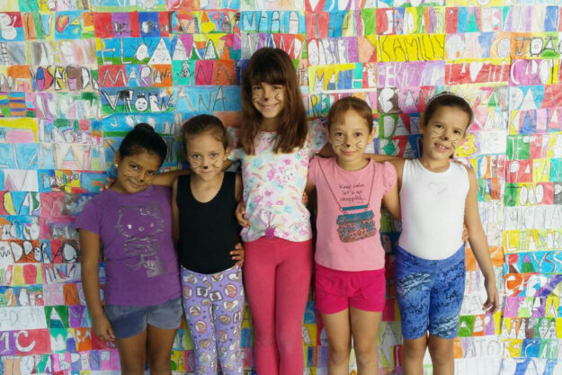 Festa a fantasia: Grupo de meninas com maquiagem de gatinho em frente a um mural de arte. Cinco meninas, com maquiagem de gatinho no rosto, posam em frente a uma parede de desenhos coloridos com nomes.