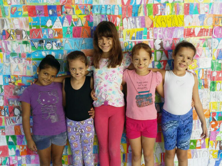 Festa a fantasia: Grupo de meninas com maquiagem de gatinho em frente a um mural de arte. Cinco meninas, com maquiagem de gatinho no rosto, posam em frente a uma parede de desenhos coloridos com nomes.