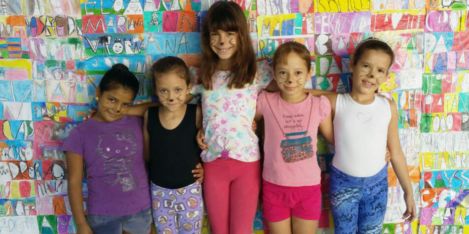 Festa a fantasia: Grupo de meninas com maquiagem de gatinho em frente a um mural de arte. Cinco meninas, com maquiagem de gatinho no rosto, posam em frente a uma parede de desenhos coloridos com nomes.