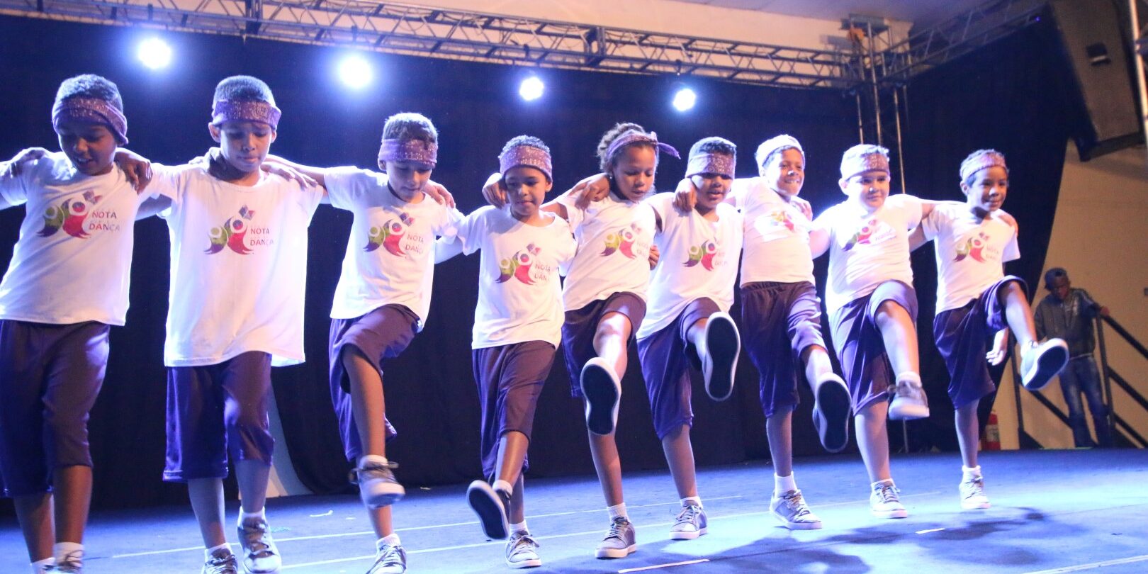 Apresentação de dança em grupo de meninos com coreografia sincronizada. Oito meninos, com bandanas e uniformes brancos e roxos, dançam com as mãos nos ombros uns dos outros em um palco iluminado.