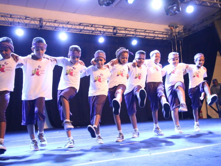 Apresentação de dança em grupo de meninos com coreografia sincronizada. Oito meninos, com bandanas e uniformes brancos e roxos, dançam com as mãos nos ombros uns dos outros em um palco iluminado.
