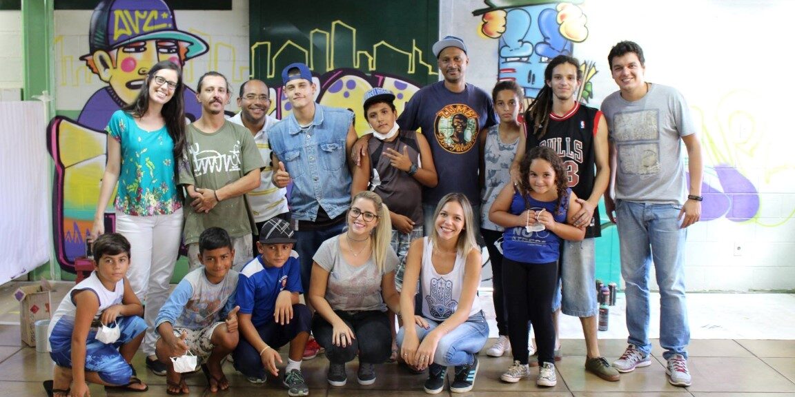 Encontro de grupo em sala com arte urbana. Grupo de adultos e crianças posa para foto em frente a uma parede de escola com grafite.