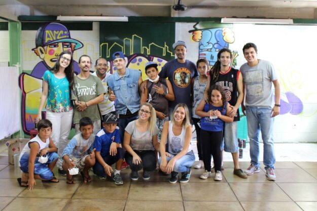 Encontro de grupo em sala com arte urbana. Grupo de adultos e crianças posa para foto em frente a uma parede de escola com grafite.