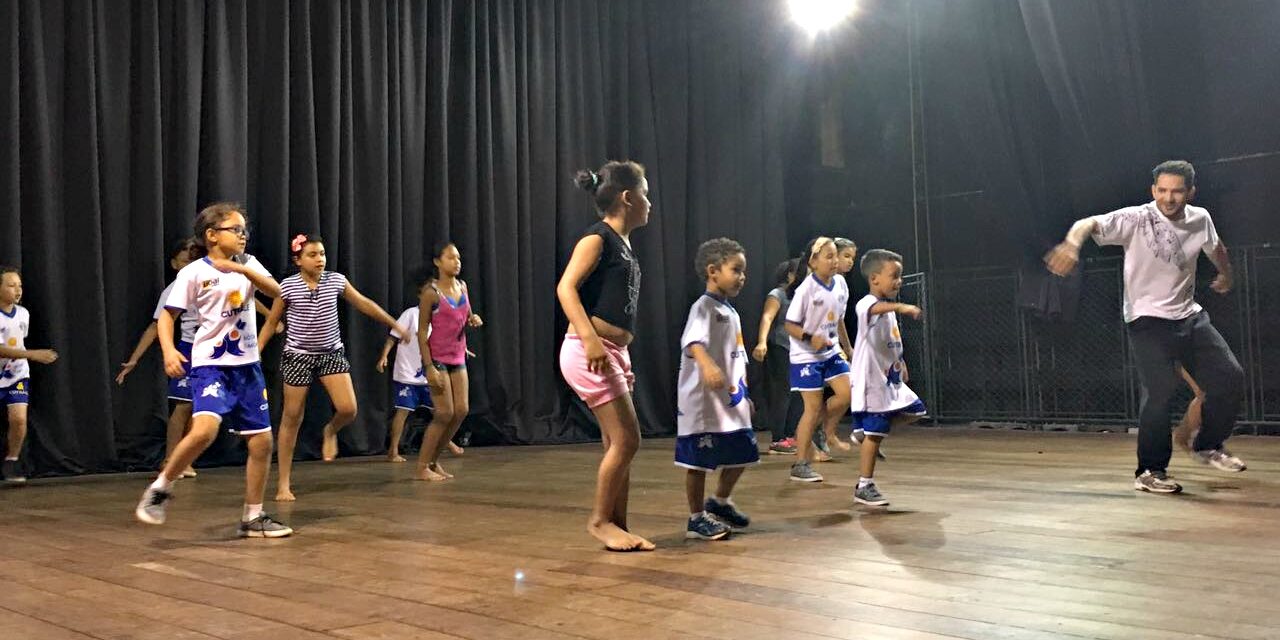 Ensaio de dança: crianças e professor praticam coreografia no palco. Adulto e crianças em trajes casuais praticam movimentos de dança em um palco de madeira com cortinas pretas no fundo.