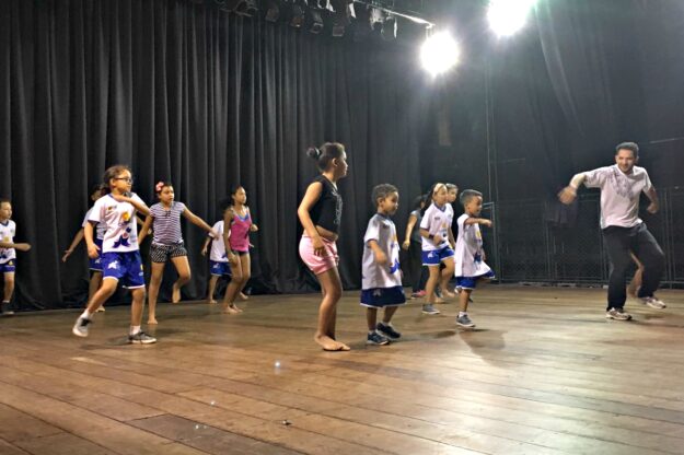 Ensaio de dança: crianças e professor praticam coreografia no palco. Adulto e crianças em trajes casuais praticam movimentos de dança em um palco de madeira com cortinas pretas no fundo.