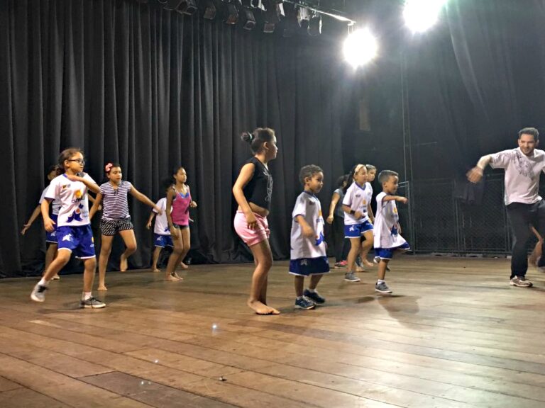 Ensaio de dança: crianças e professor praticam coreografia no palco. Adulto e crianças em trajes casuais praticam movimentos de dança em um palco de madeira com cortinas pretas no fundo.