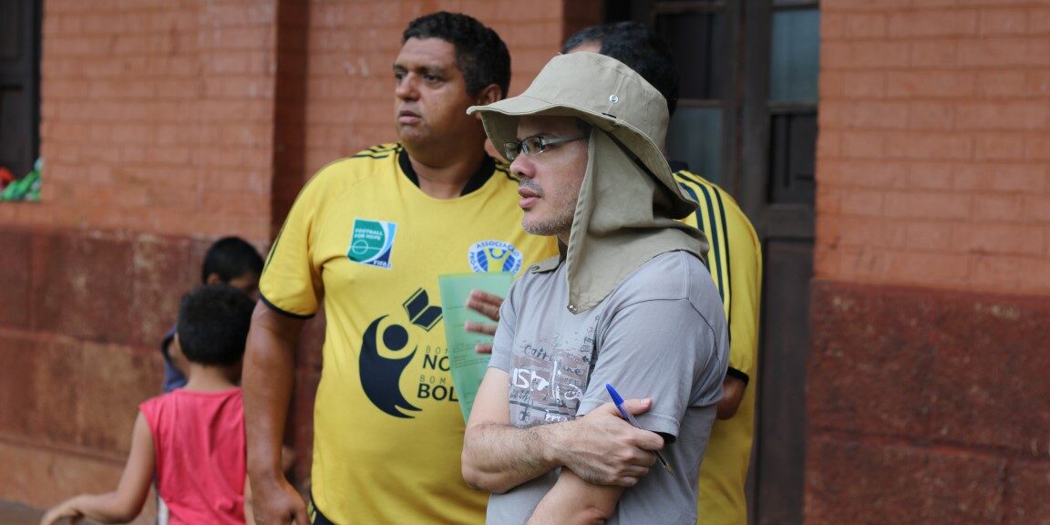 Coordenadores e apoiadores observam o evento esportivo. Dois homens, um de chapéu e camisa cinza e outro com camisa amarela "Nota 10 na Bola", observam uma atividade ao ar livre.