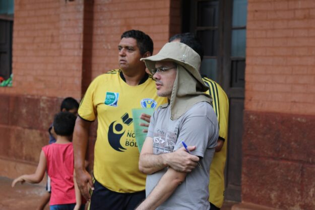 Dois homens, um de chapéu e camisa cinza e outro com camisa amarela "Nota 10 na Bola", observam uma atividade ao ar livre.
