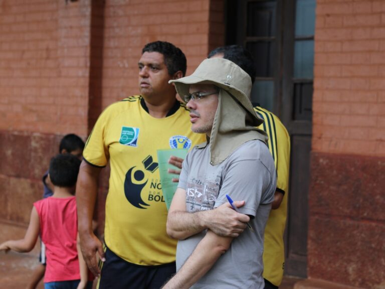 Coordenadores e apoiadores observam o evento esportivo. Dois homens, um de chapéu e camisa cinza e outro com camisa amarela "Nota 10 na Bola", observam uma atividade ao ar livre.