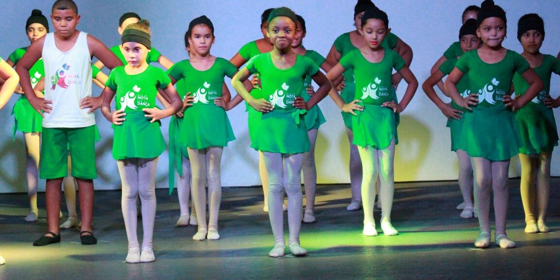 Crianças em figurino verde se apresentam no palco. Grupo de crianças e adolescentes em trajes de balé e dança verdes no palco, com as mãos na cintura e olhando para frente.