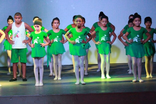 Crianças em figurino verde se apresentam no palco. Grupo de crianças e adolescentes em trajes de balé e dança verdes no palco, com as mãos na cintura e olhando para frente.