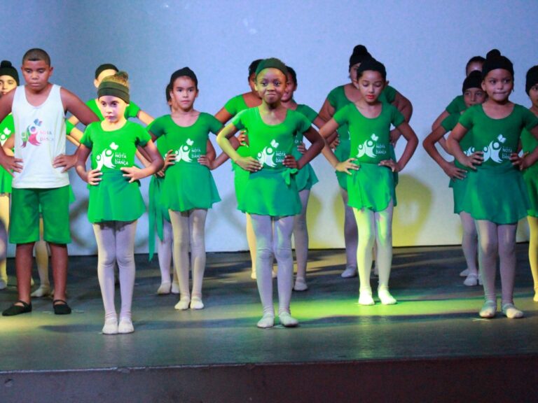 Crianças em figurino verde se apresentam no palco. Grupo de crianças e adolescentes em trajes de balé e dança verdes no palco, com as mãos na cintura e olhando para frente.