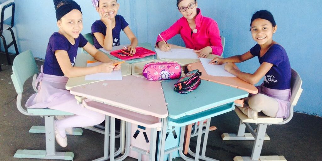 Estudantes de balé em atividade de desenho na sala de aula. Crianças vestidas para balé, sentadas em uma mesa hexagonal em uma sala de aula, sorriem enquanto desenham.