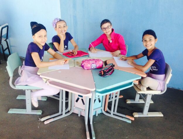 Estudantes de balé em atividade de desenho na sala de aula. Crianças vestidas para balé, sentadas em uma mesa hexagonal em uma sala de aula, sorriem enquanto desenham.
