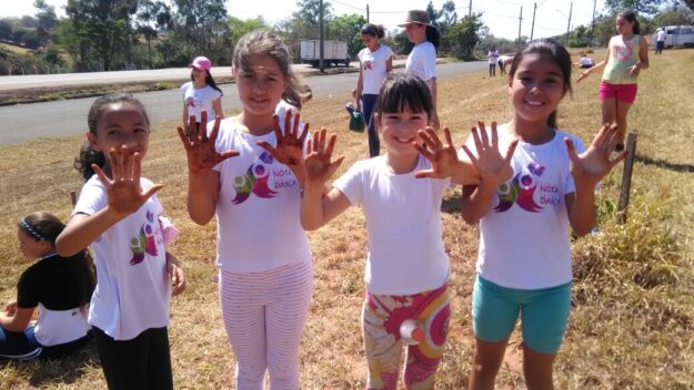 Crianças mostram as mãos sujas durante atividade ao ar livre. Quatro meninas em camisetas brancas mostram as mãos cobertas de terra ou tinta, sorrindo em uma área externa gramada.