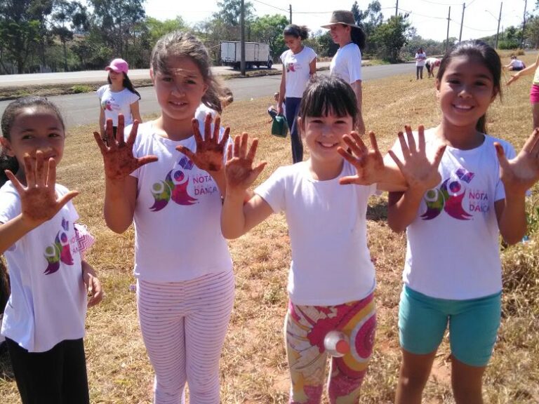 Crianças mostram as mãos sujas durante atividade ao ar livre. Quatro meninas em camisetas brancas mostram as mãos cobertas de terra ou tinta, sorrindo em uma área externa gramada.