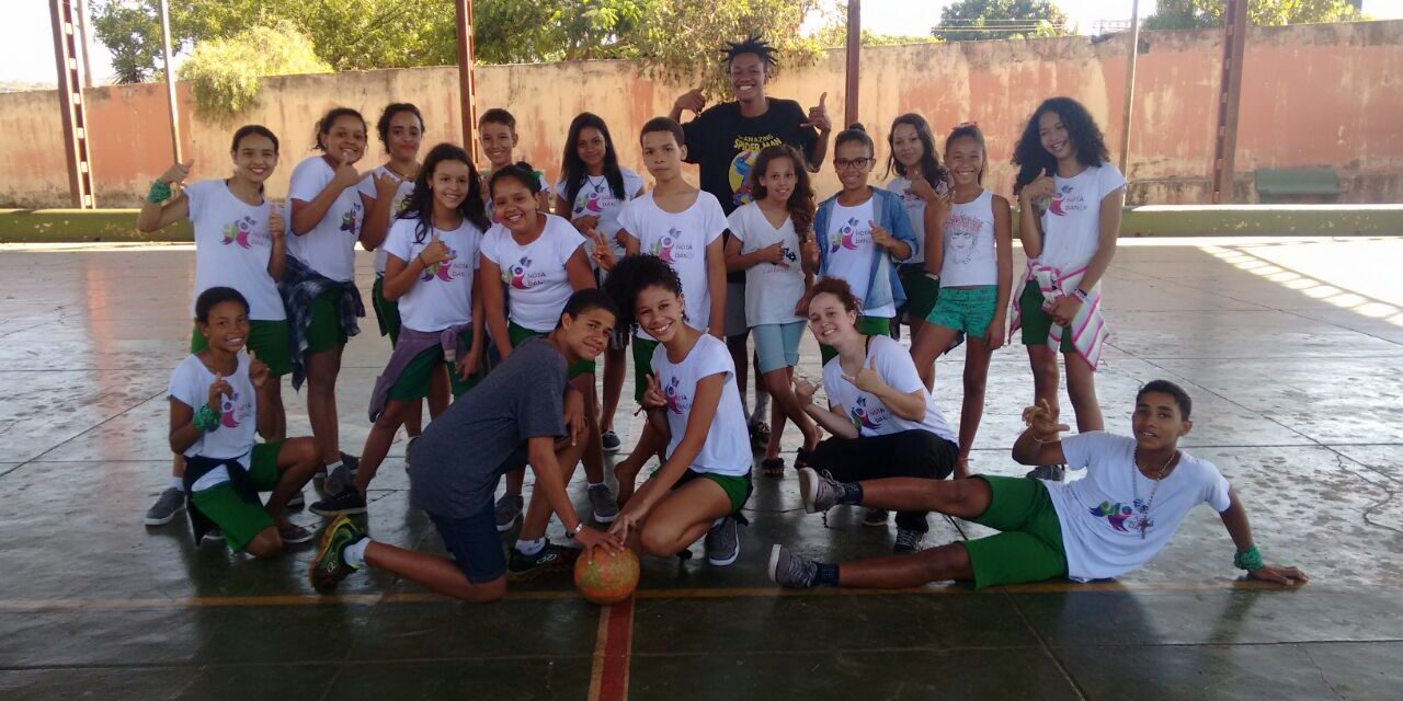 Foto de grupo de jovens em quadra com bola. Grupo de crianças em camisetas brancas e shorts verdes posa em uma quadra, alguns com polegares para cima, com uma bola no centro.